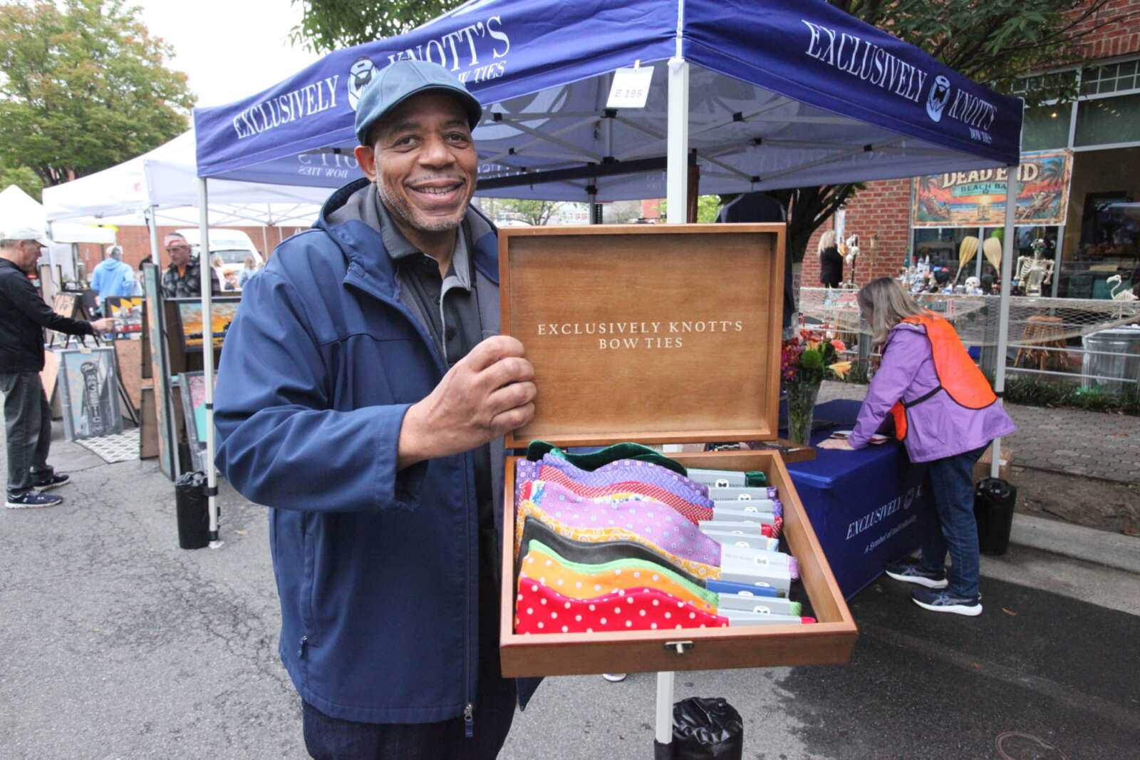 Smiling man in a blue jacket holds an open wooden box displaying colorful bow ties at an outdoor market stall.