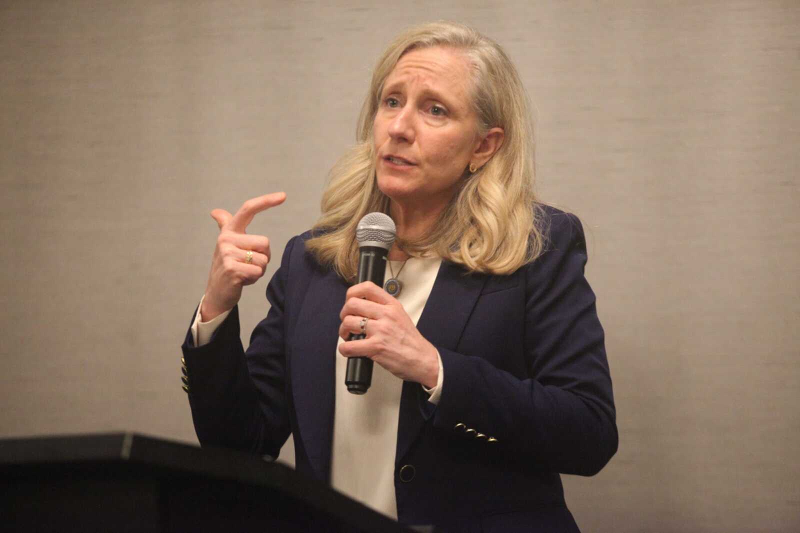 Blonde woman in a navy blazer speaks into a handheld microphone, gesturing with her right hand while presenting.