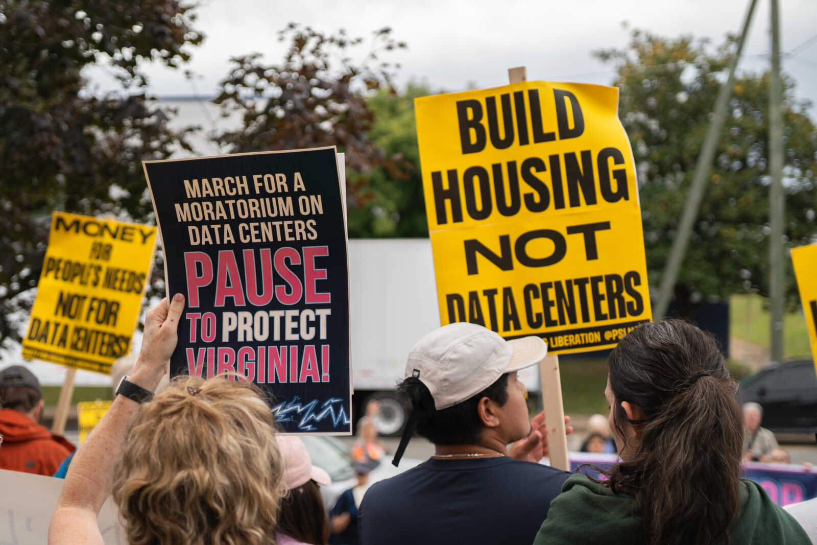 Crowd of protesters holding signs, including a yellow sign that says "BUILD HOUSING NOT DATA CENTERS" and a dark sign reading "PAUSE TO PROTECT VIRGINIA!"