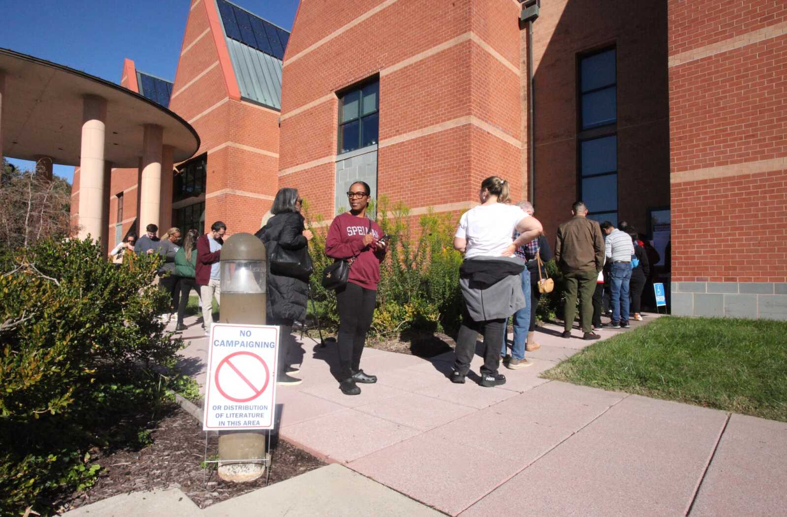 People line up outside a brick campus building on a sunny day; a 'No campaigning' sign is in the foreground near a lamp post.