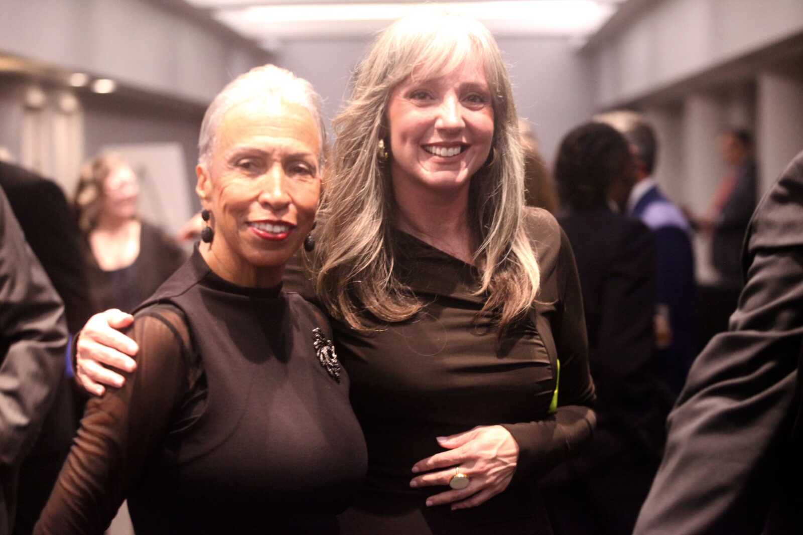 Two women in black dresses pose together at a social event, smiling at the camera.