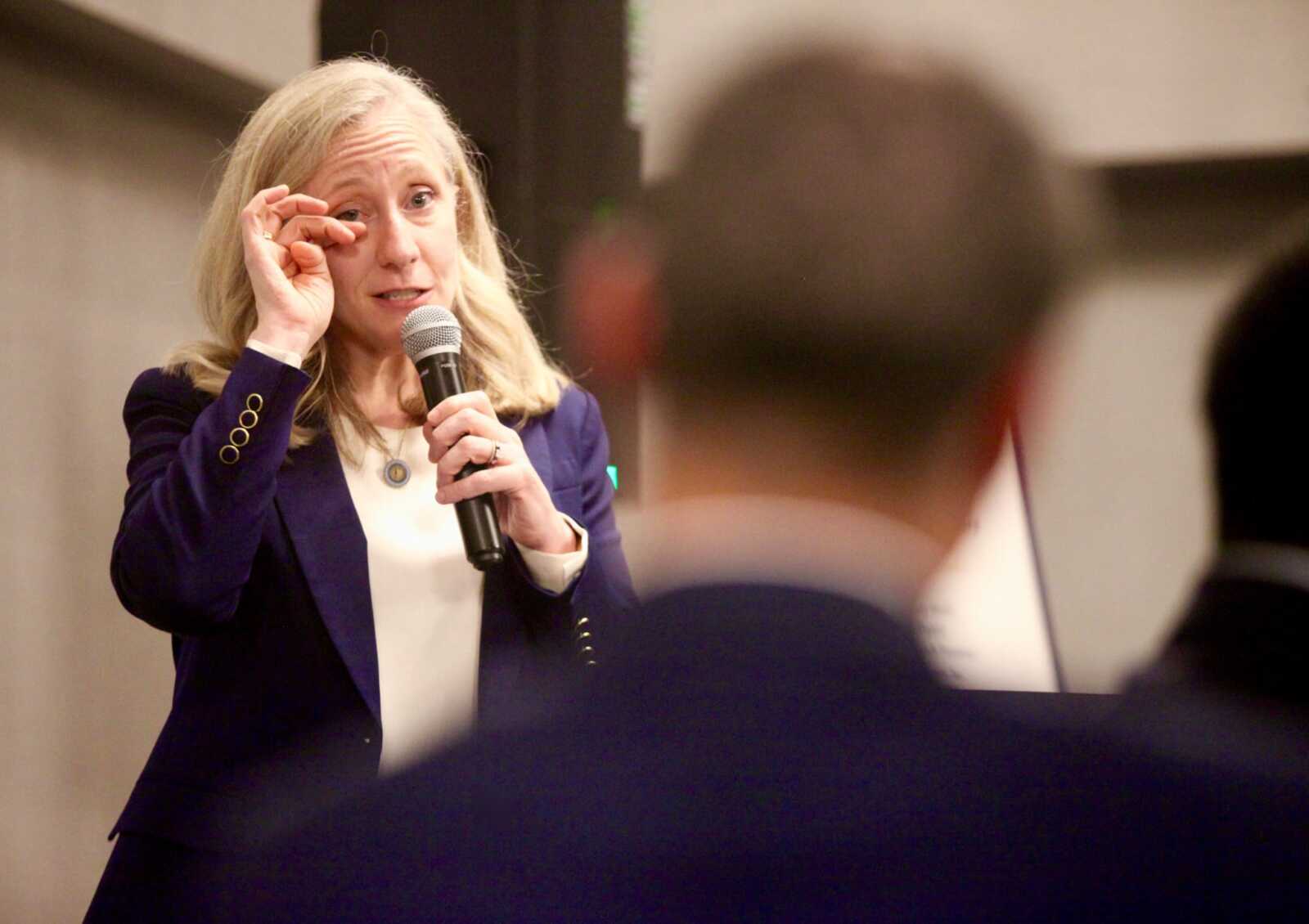 Woman with blonde hair in a purple blazer speaks into a handheld microphone to an audience, gesturing with her hand near her eye.