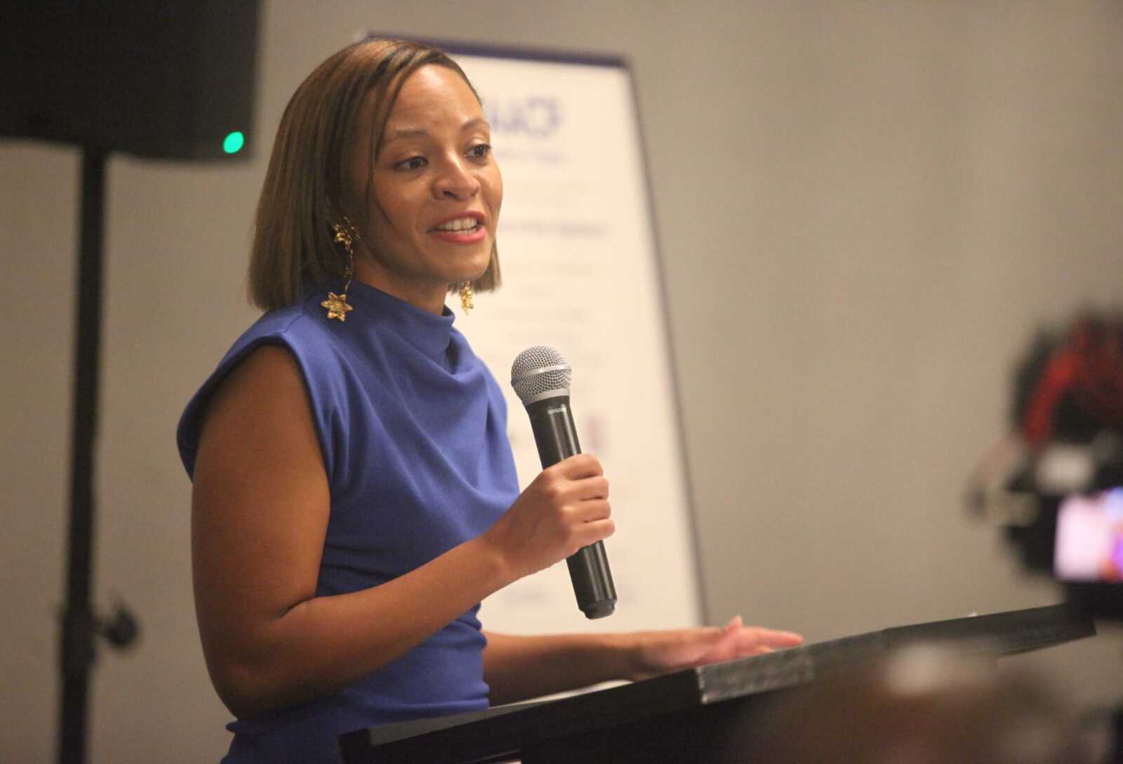 Woman in a blue sleeveless dress speaks into a handheld microphone at a podium, delivering a talk.