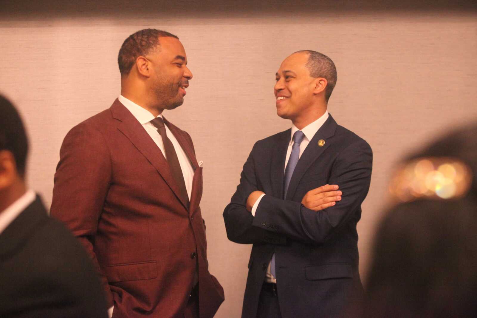 Two men in suits smiling at each other during a formal event, with blurred audience in the foreground.