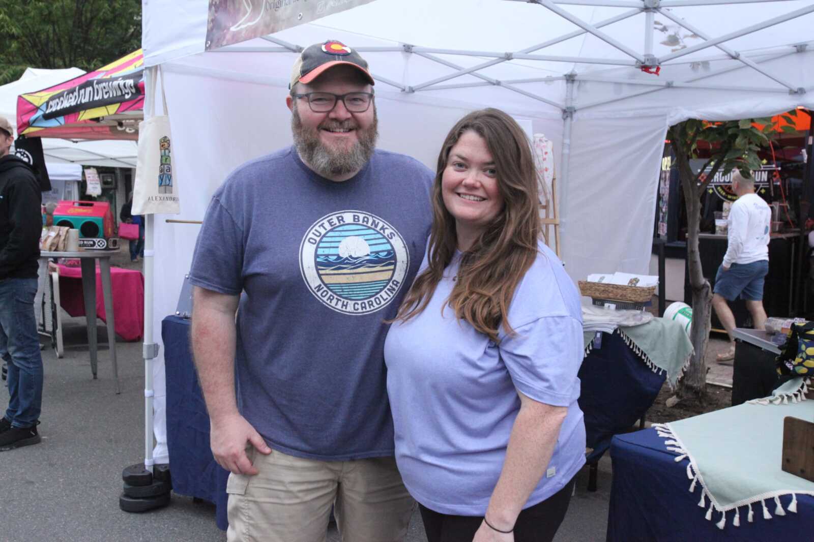 Two smiling adults pose together at an outdoor market with white canopy tents in the background.