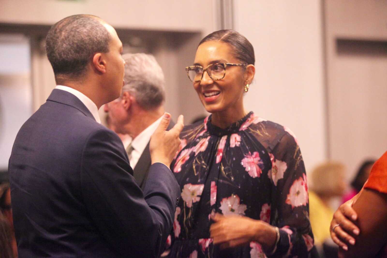 Man in a navy suit speaks with a smiling woman wearing a floral blouse and glasses at a social event.