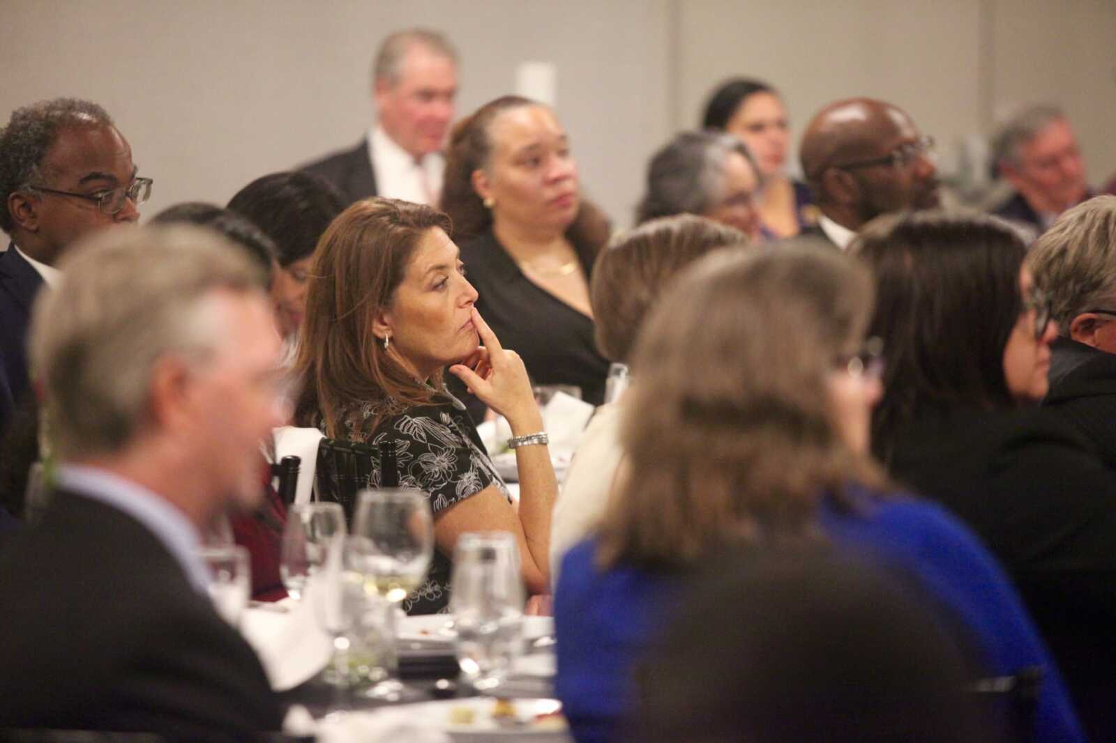A woman in a black floral dress sits at a table during a formal event, resting her chin on her hand as she listens.