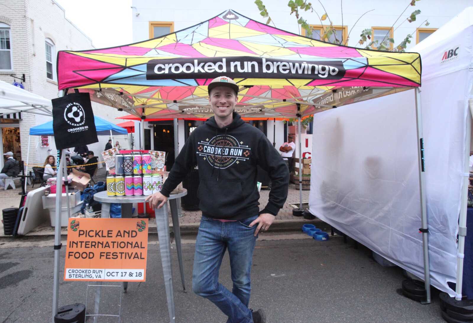 A smiling man in a black Crooked Run hoodie stands under a colorful festival tent with a sign for Pickle and International Food Festival nearby.