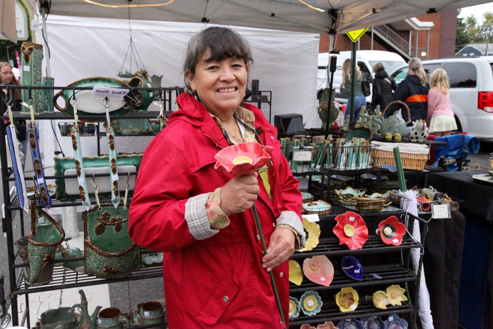 Smiling woman in a red jacket holds a pink ceramic flower-shaped bowl at a pottery stall.