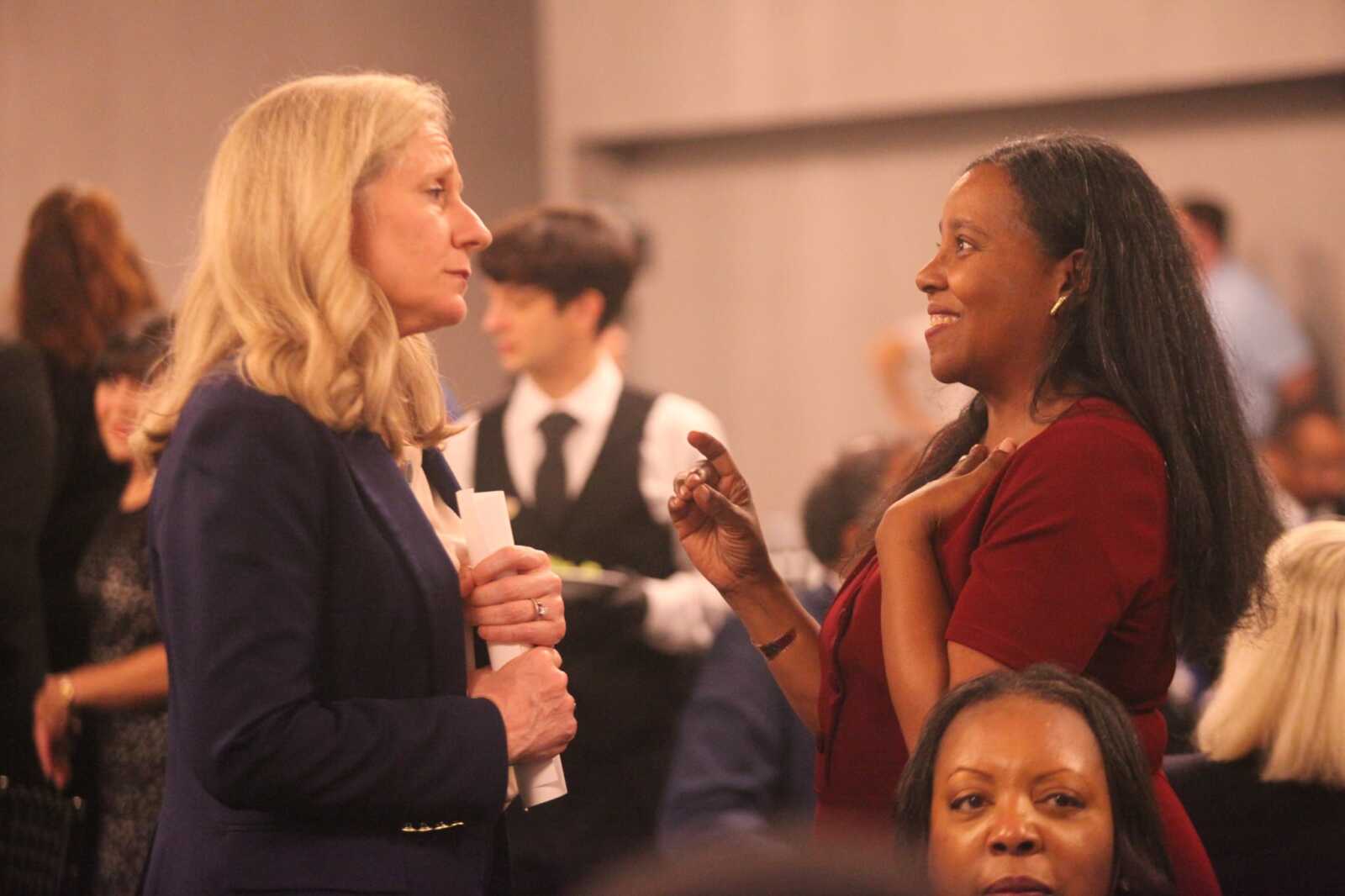 Two women converse at a formal event: a blonde in a navy blazer holds rolled papers while a woman in red smiles and gestures nearby.