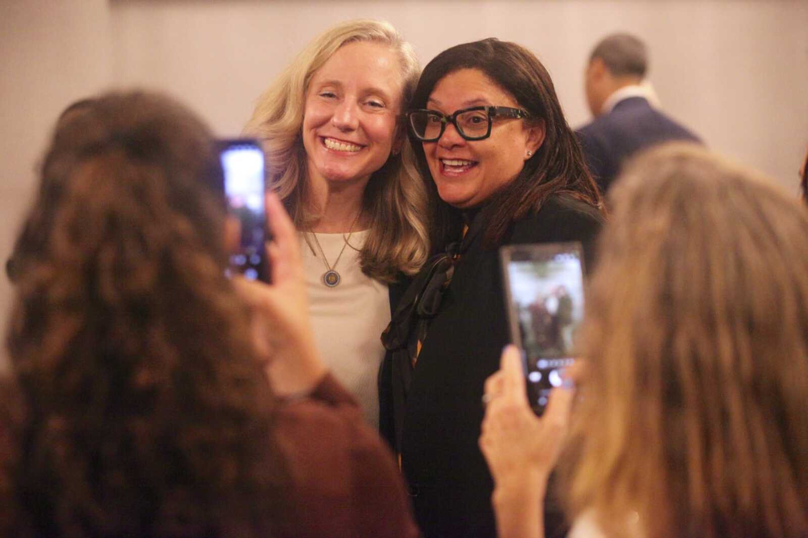 Two women posing for a photo at a crowded event; one blond in white, the other dark-haired with glasses.