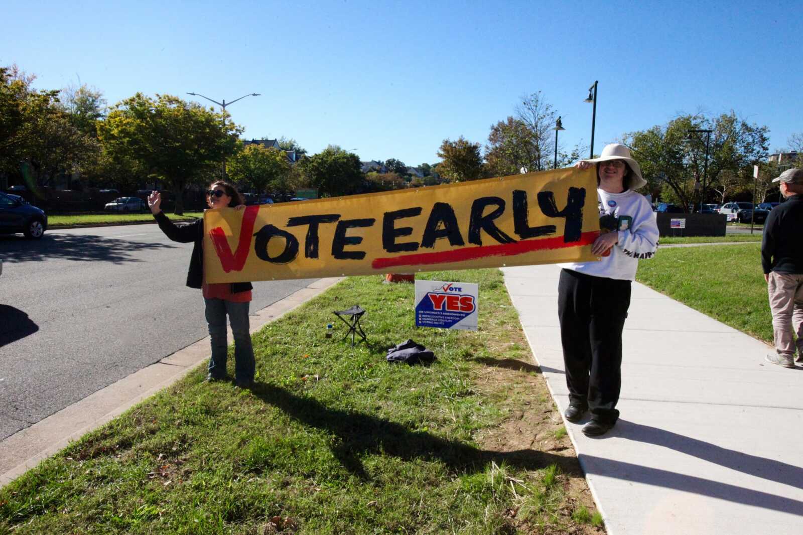 Two people stand on a sunny curb holding a large yellow banner that reads 'VOTE EARLY' across the frame behind a sidewalk.