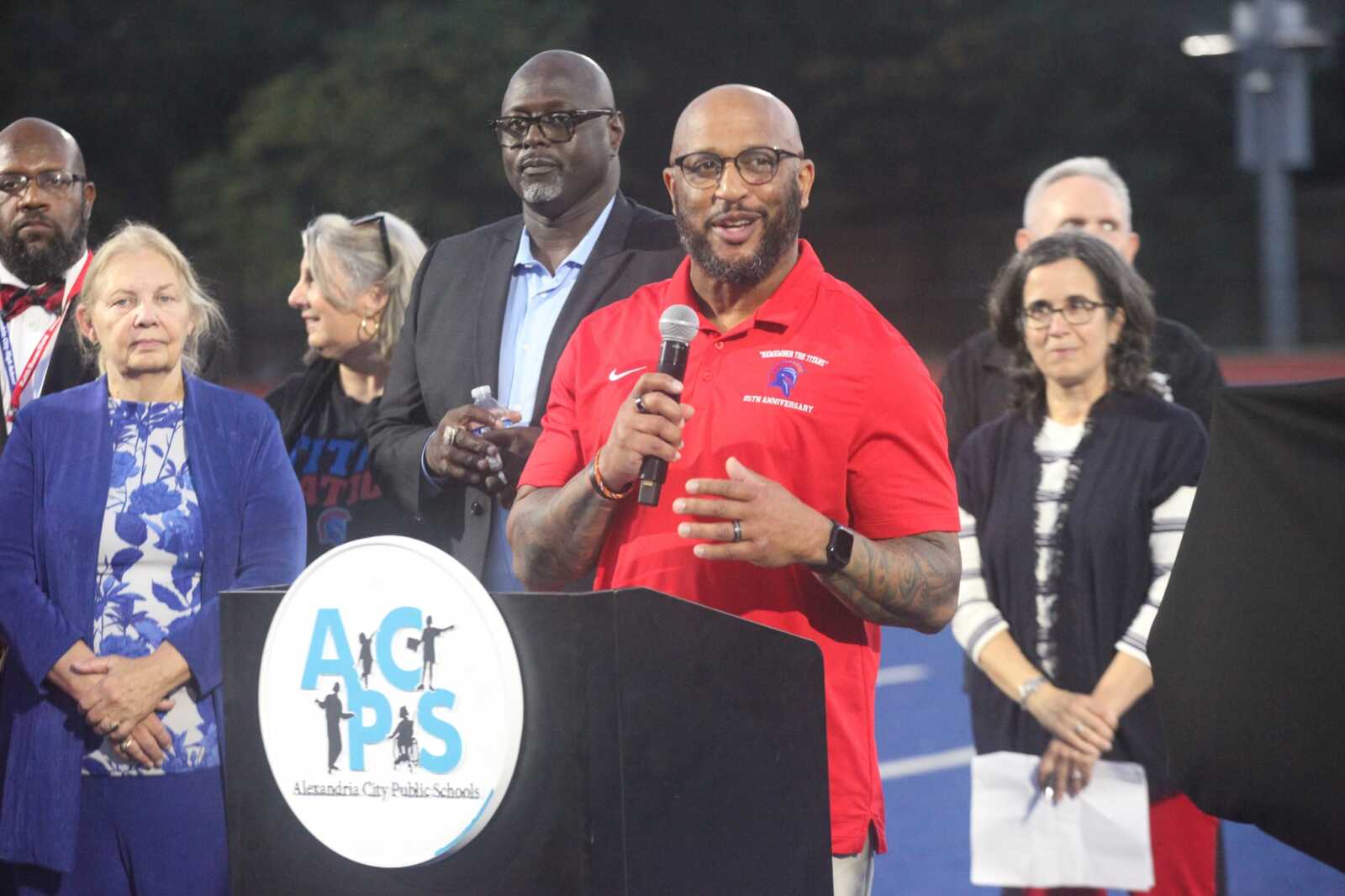 Man in a red polo speaks into a microphone at a podium with a circular logo, while several adults stand behind him on a track field during a school event.