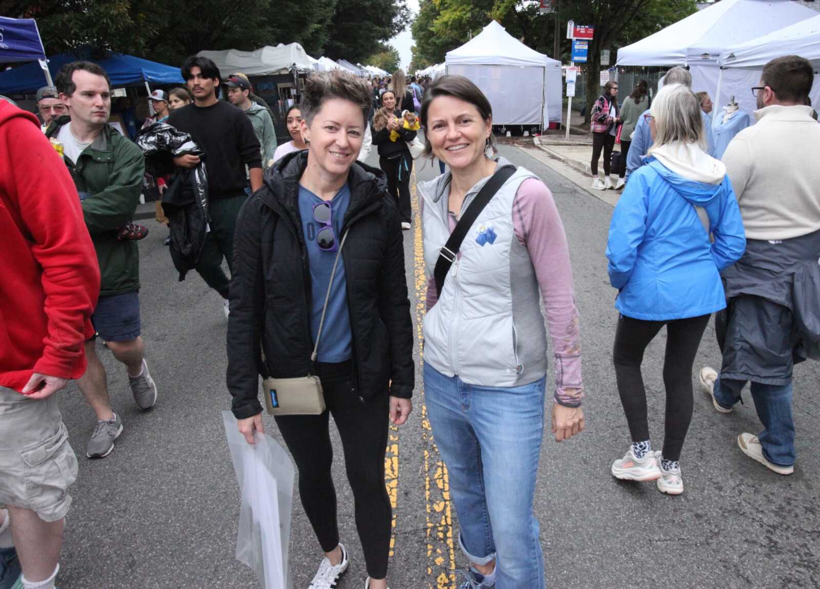 Two women stand smiling on a street fair, with tents and people in the background.
