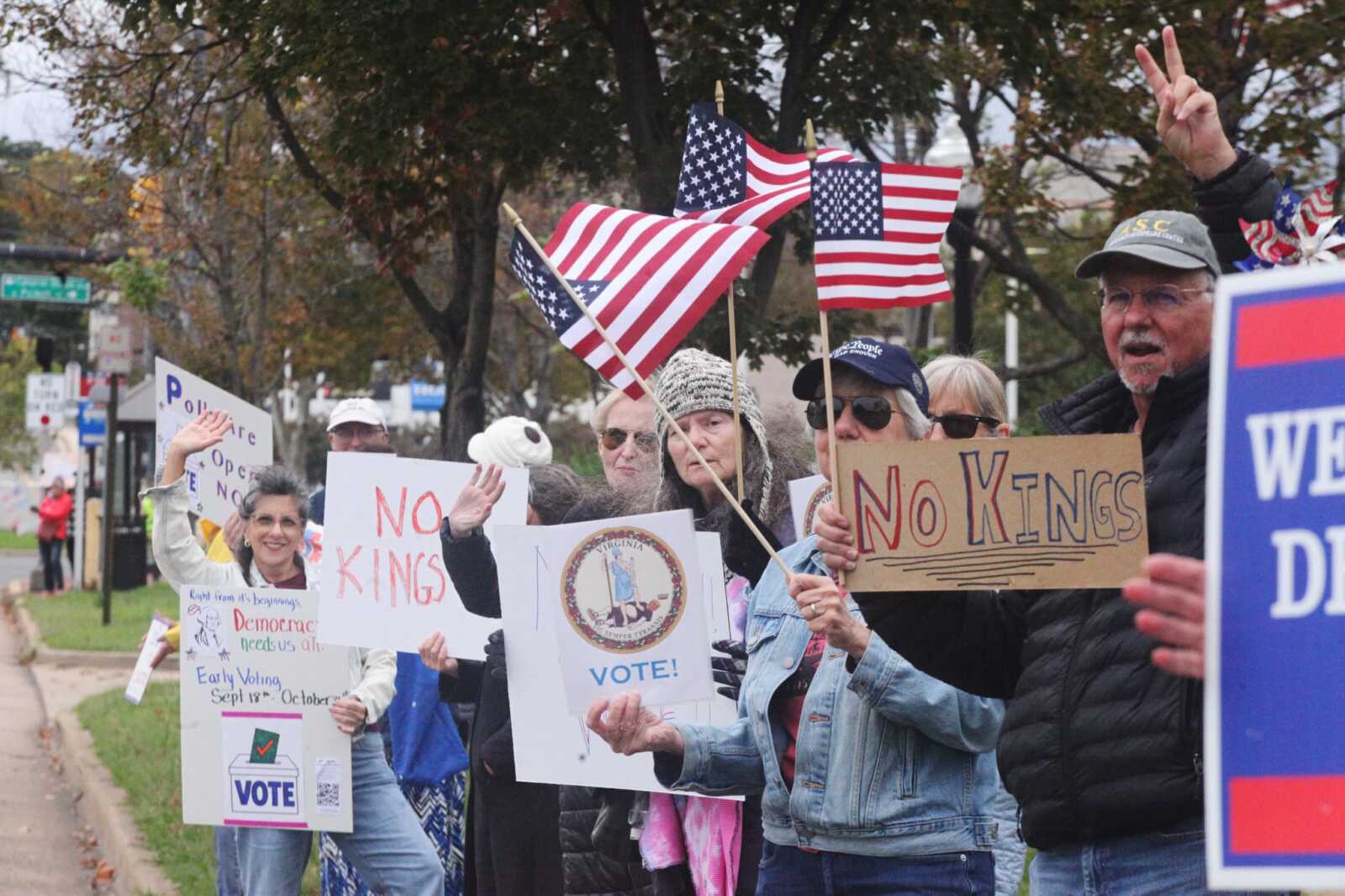 Crowd of protesters holding No Kings sign and American flags at a street rally; Virginia state seal visible on one sign.