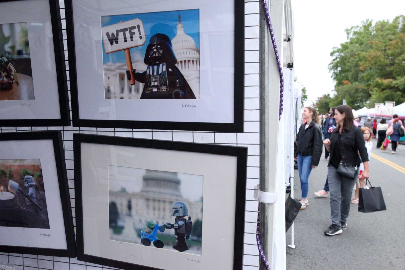 Framed LEGO minifigure art on a grid display at an outdoor festival, with shoppers walking along a street in the background.