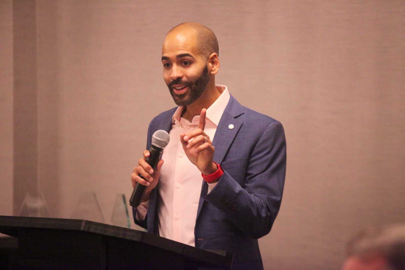 Man in a blue blazer and pink shirt speaks at a podium, holding a microphone and gesturing with his finger raised.