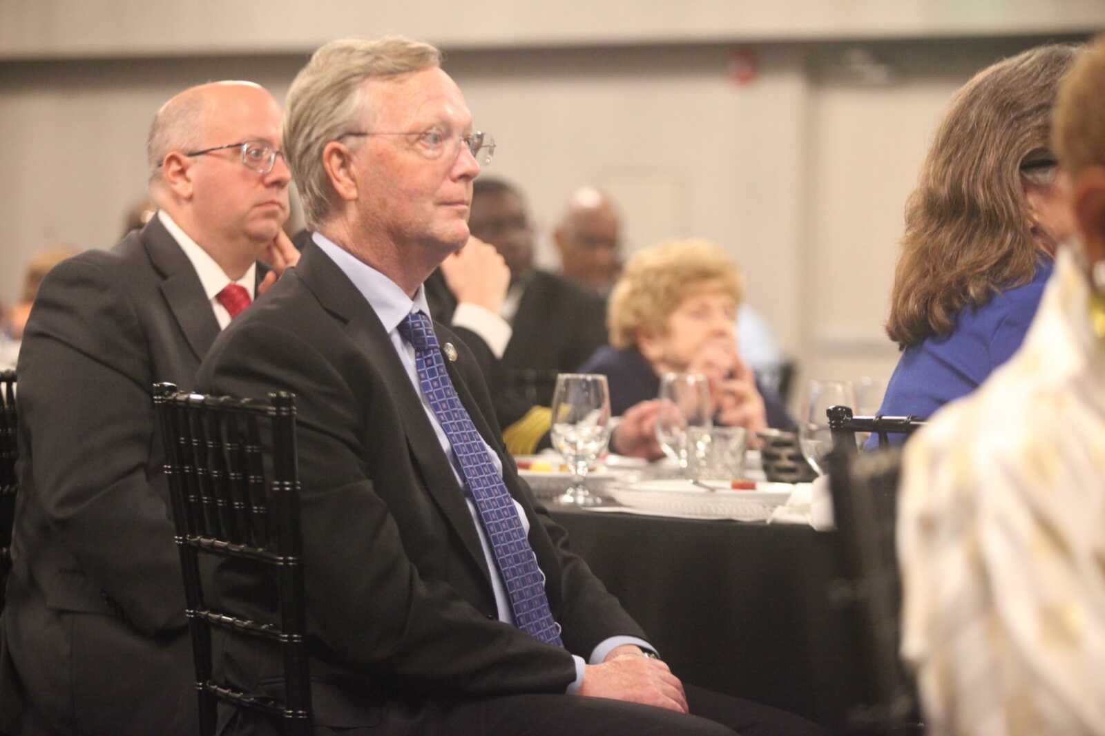 Man in a dark suit and purple patterned tie sits at a formal dinner event, looking to the right; glassware and table settings in front.