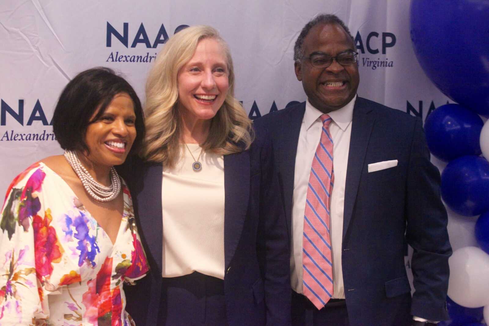Three smiling adults stand together at a civic event, with a backdrop reading NAACP Alexandria; balloons on the right. Left: woman in a floral dress and pearl necklace; center: woman in white top and navy blazer; right: man in a suit with pink striped tie.