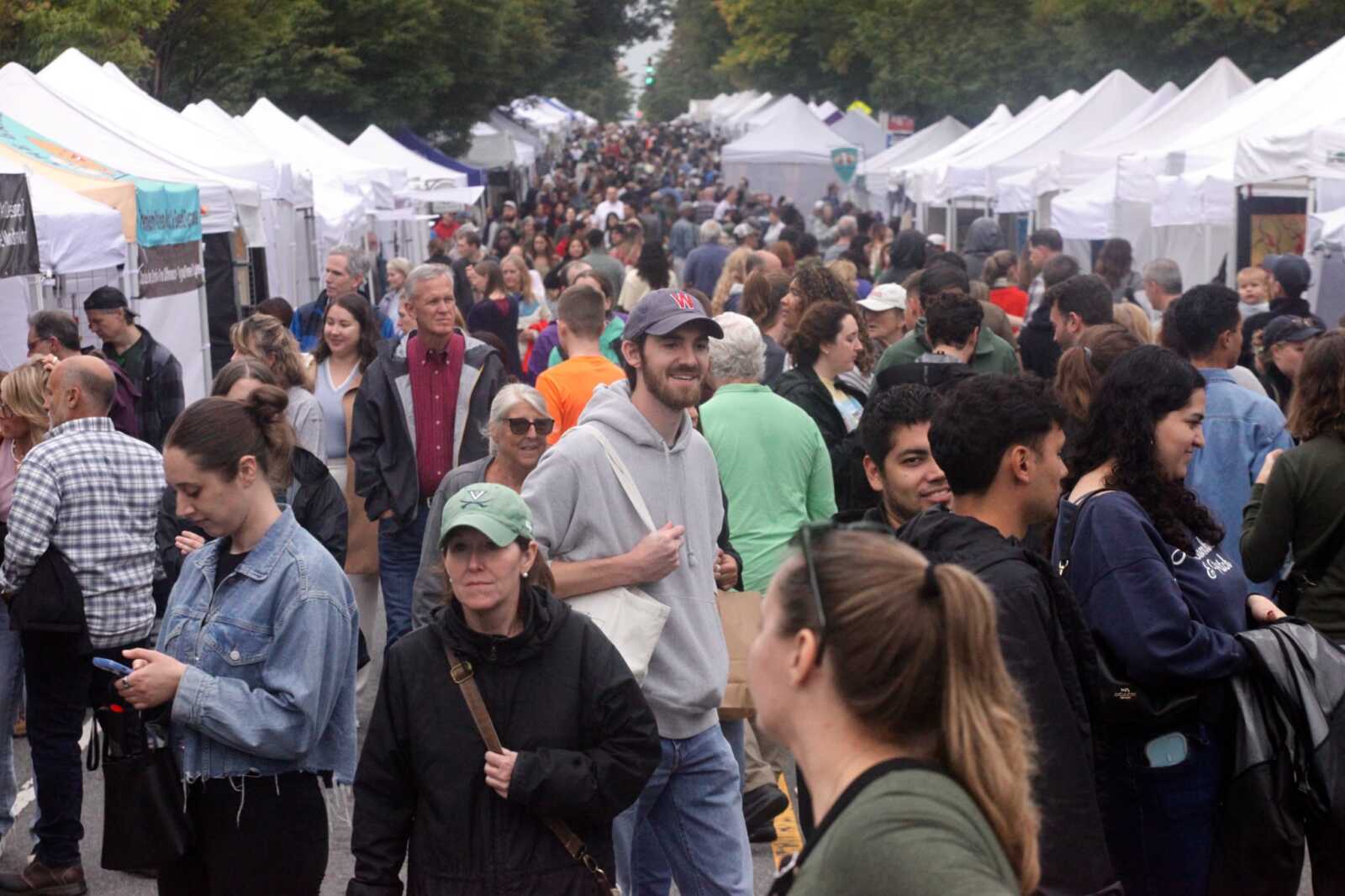 Crowded outdoor market street with white vendor tents on both sides and many shoppers.