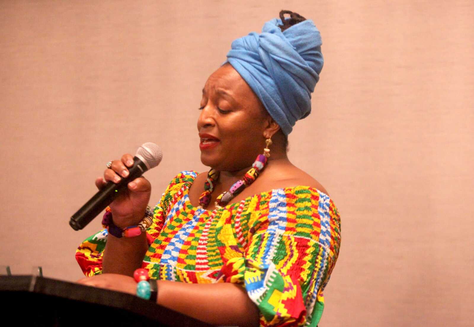 Woman wearing a blue headwrap and a colorful patterned dress sings into a microphone on stage.