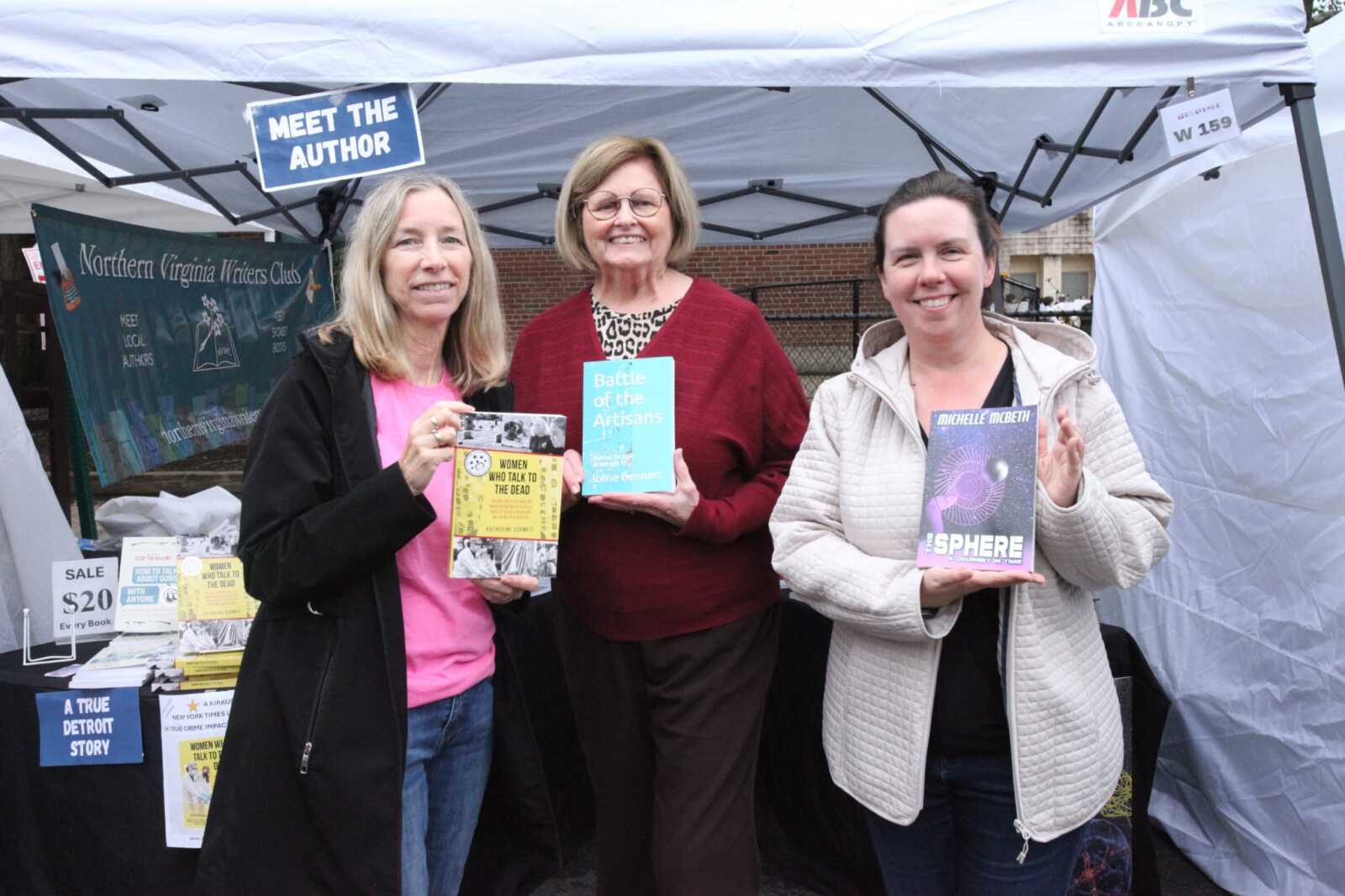 Three women at a book booth under a white tent, smiling and holding books.