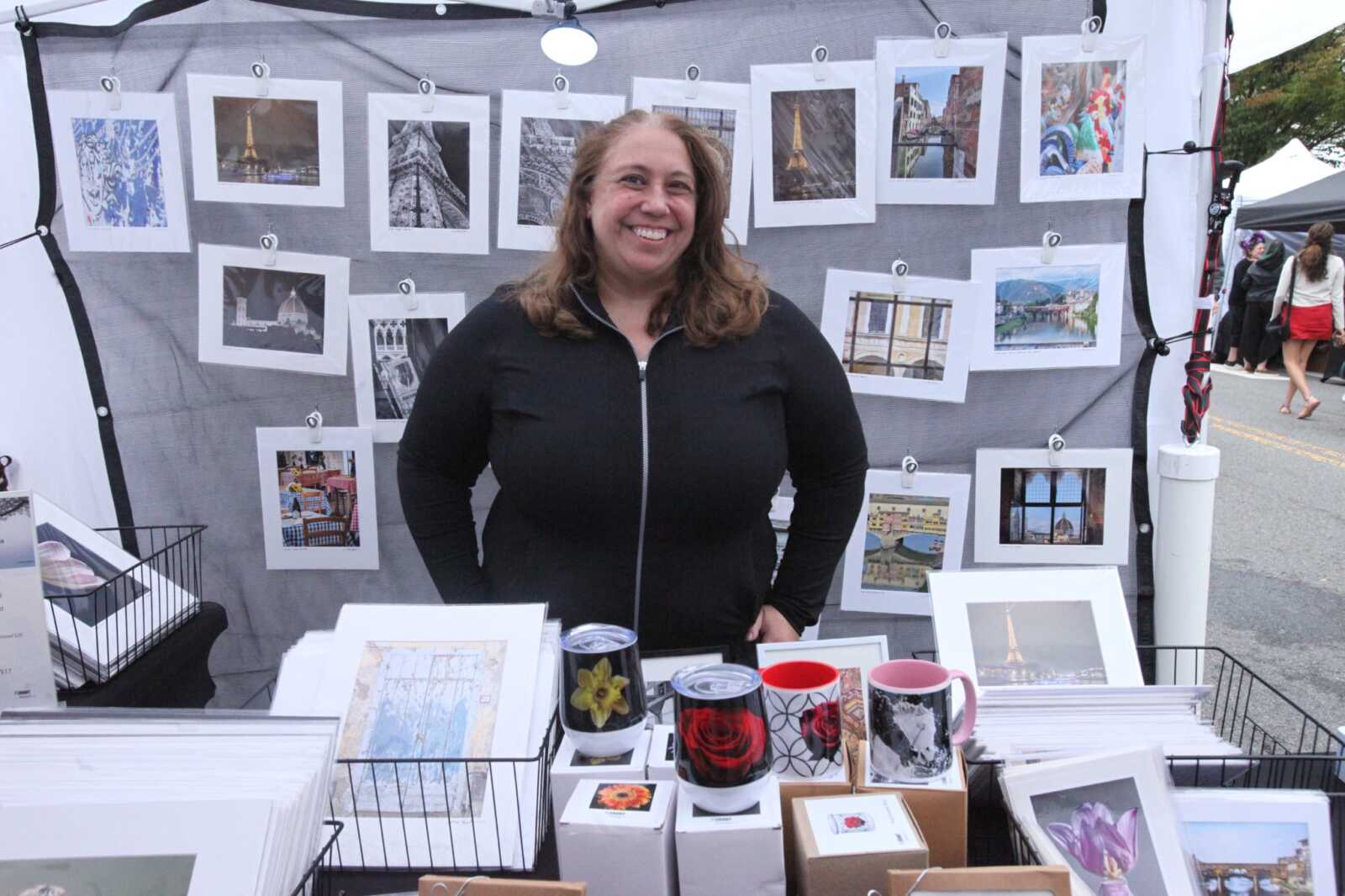Smiling woman in black zip jacket stands behind a street market stall with framed photos and mugs for sale on display shelves and hanging photos behind her
