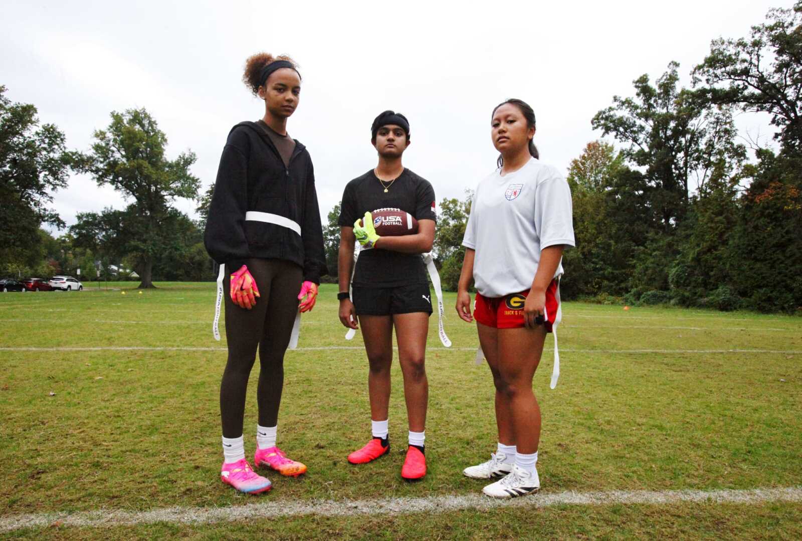 Three young women pose on a grassy field with a football, looking at the camera as teammates before a game.
