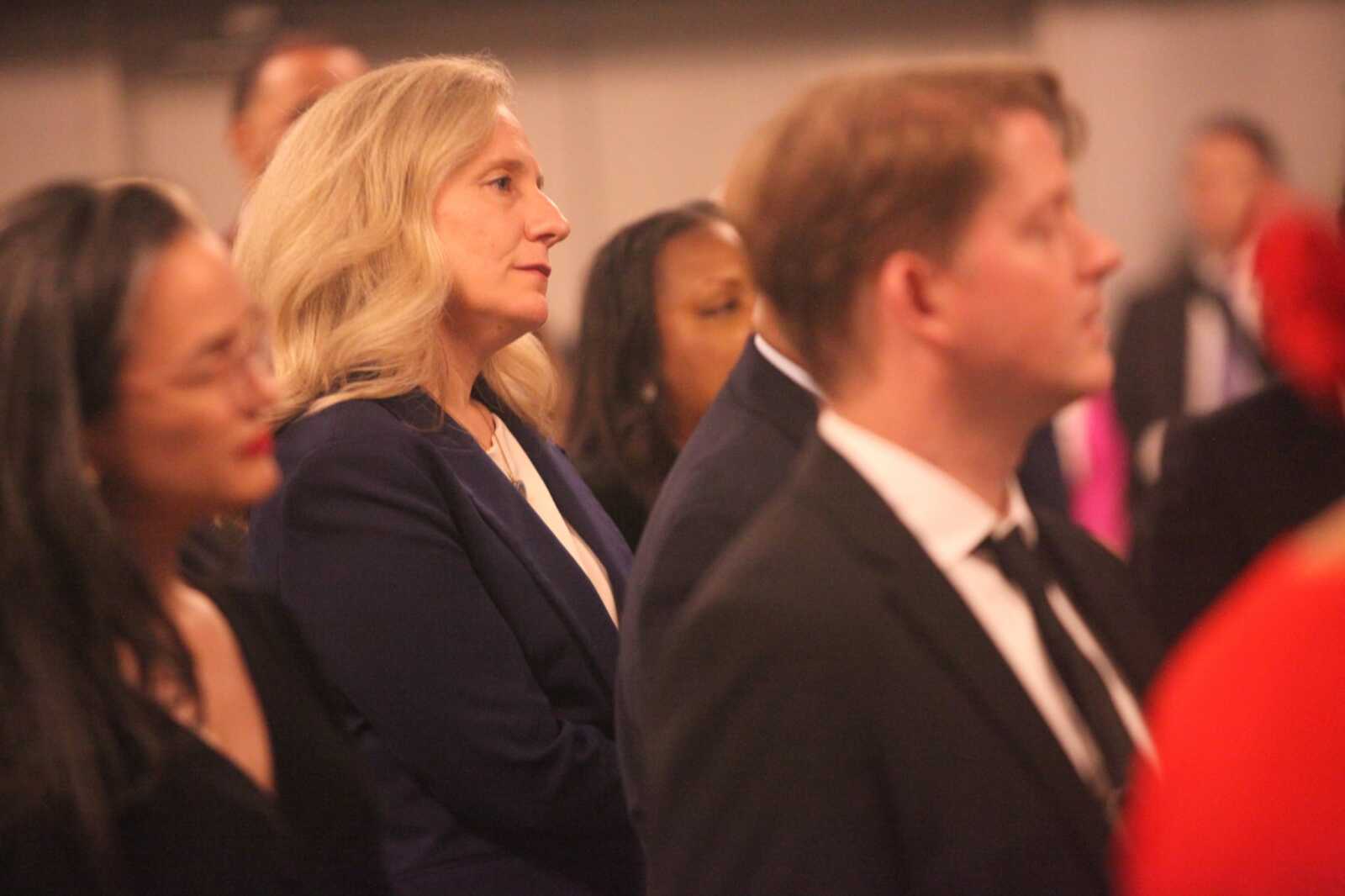 Blonde woman in a navy blazer listening intently at a formal event, surrounded by other attendees.