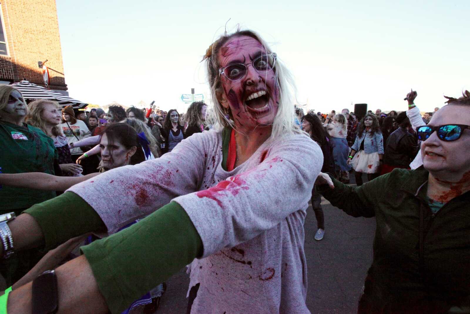 Crowd of people in zombie makeup and costumes at an outdoor event, reaching toward the camera.