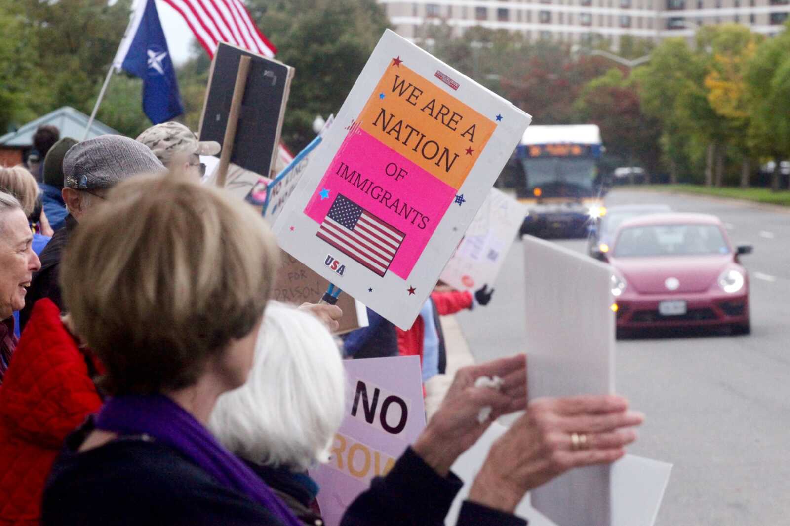 Crowd of protesters along a street, holding signs; central sign reads 'We are a nation of immigrants' with an American flag graphic