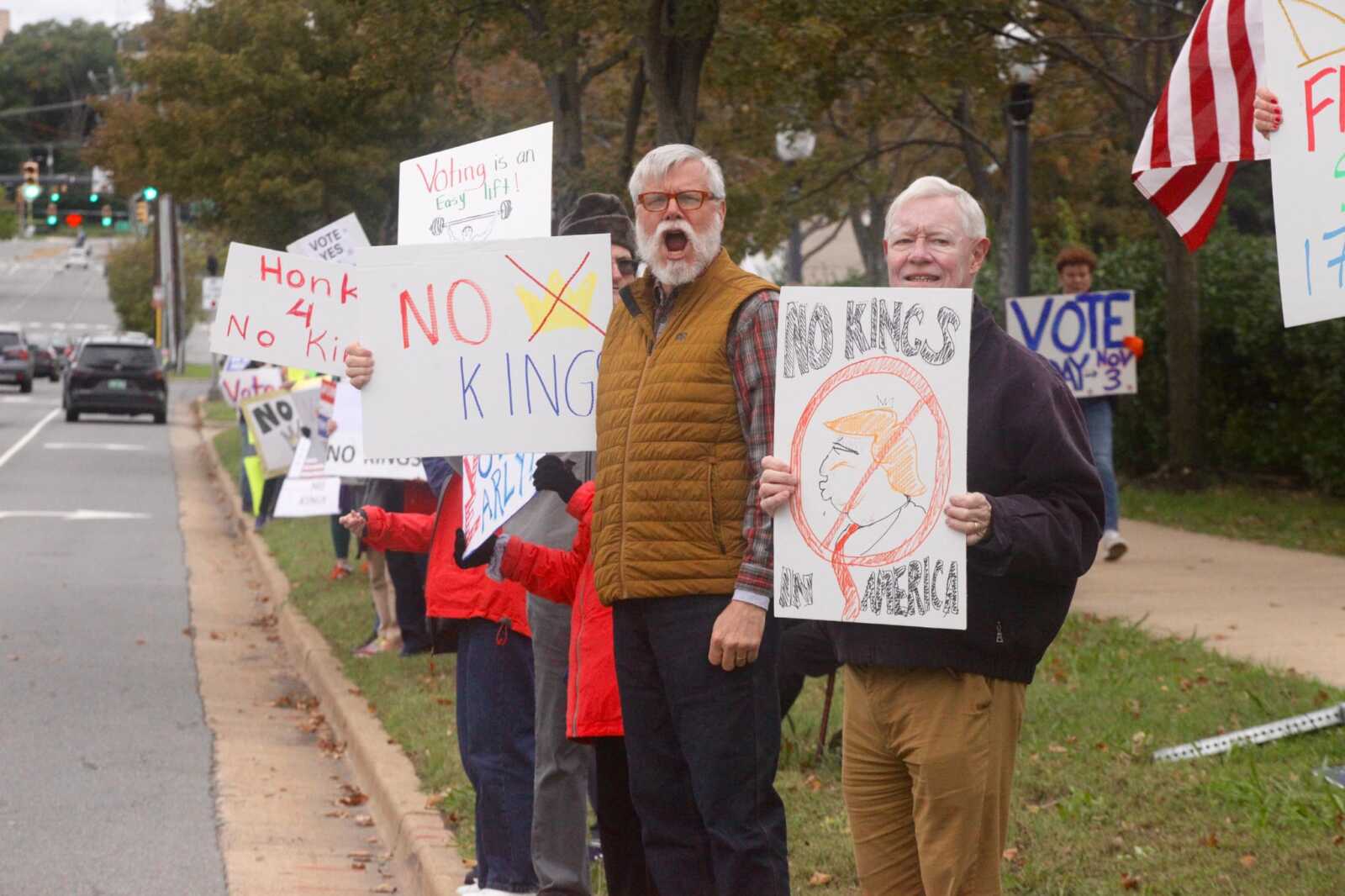A line of protesters on a sidewalk holding signs; central figure with orange vest and another man holding a 'NO KINGS' sign with a cartoon of a political figure.