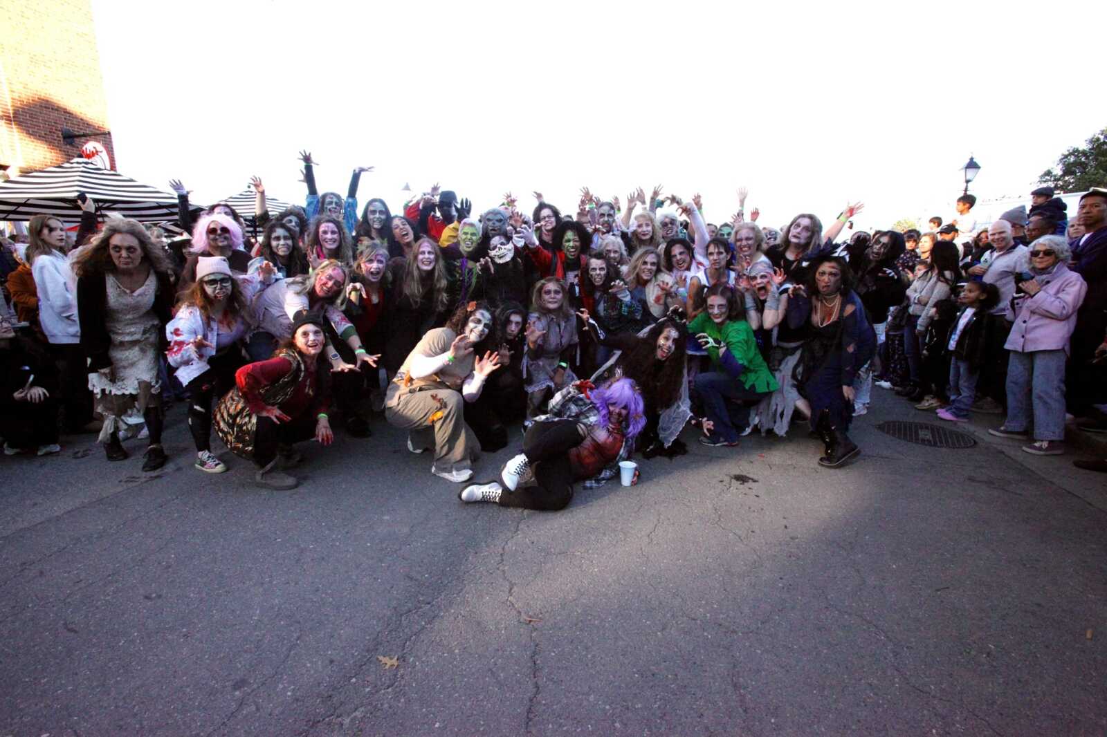 Large crowd in Halloween costumes posing as zombies on a street, many with makeup and outstretched arms, outdoors near tents and bystanders
