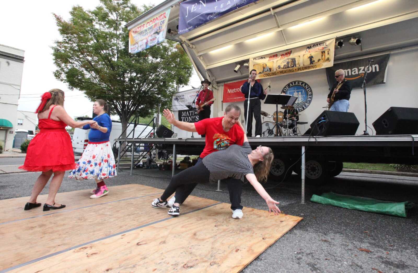 Live band performing on a stage with a dancing couple on a wooden platform in front of the crowd, outdoors at a festival.