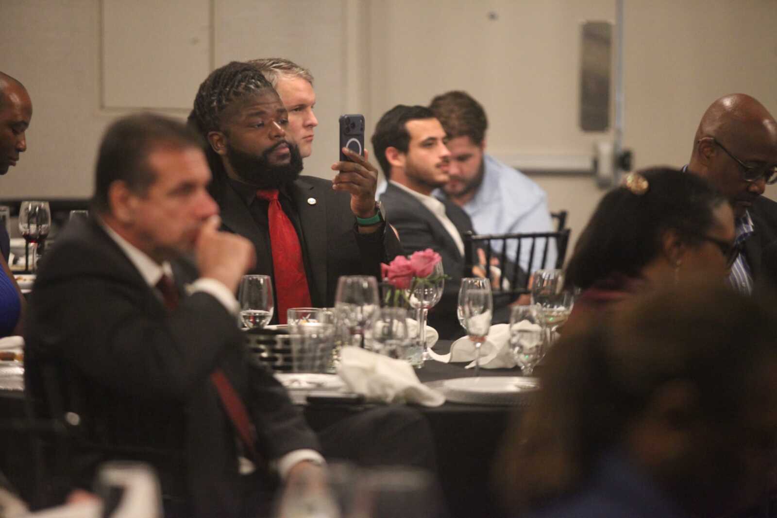 Man in a black suit with a red tie takes a selfie at a banquet table with wine glasses nearby.