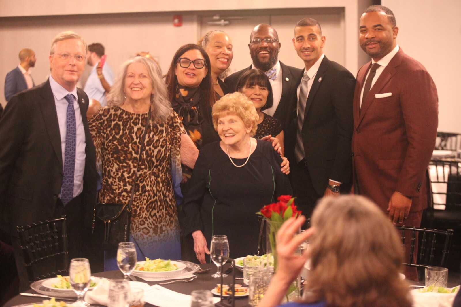 Group of eight adults posing behind a seated elderly woman at a formal banquet, smiling; table set with plates, glasses, and a red rose centerpiece.