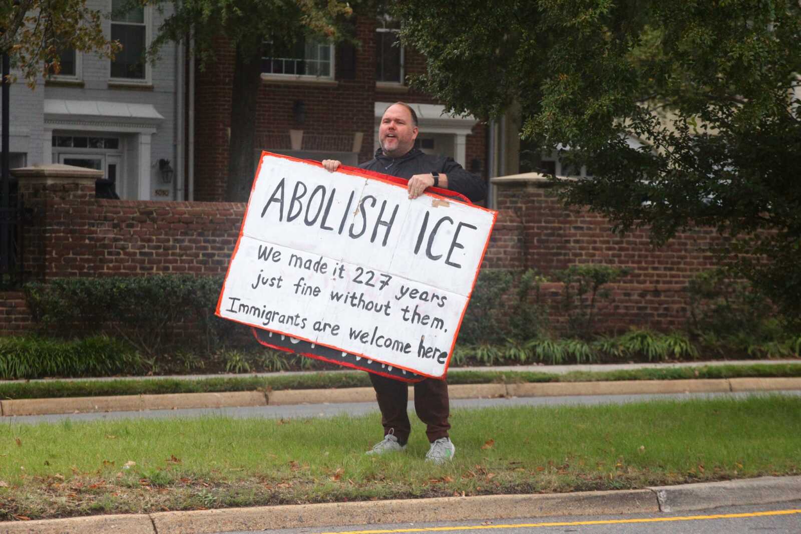 Person standing on a suburban sidewalk holding a large handwritten protest sign that says 'ABOLISH ICE' with smaller text about immigrants being welcome here, in front of brick houses and greenery.