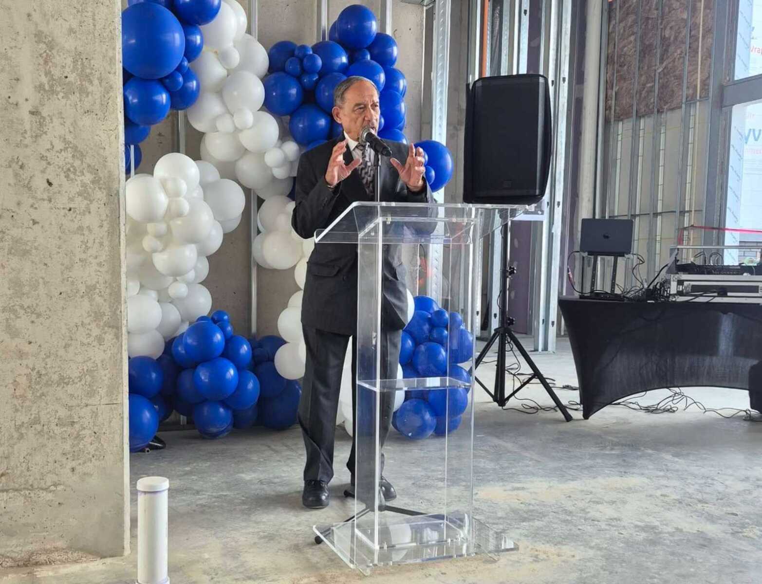 Man in suit speaking at a clear acrylic podium with a microphone in a construction-warehouse setting, blue and white balloon decorations in the background