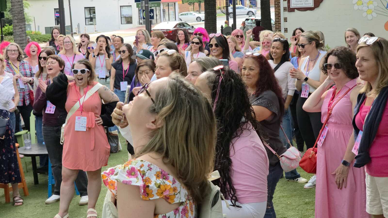 Group of smiling women at an outdoor pink-themed event, cheering together wearing lanyards and sunglasses.