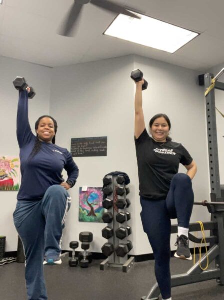 Two women in a gym performing standing overhead dumbbell presses on one leg, smiling at the camera.