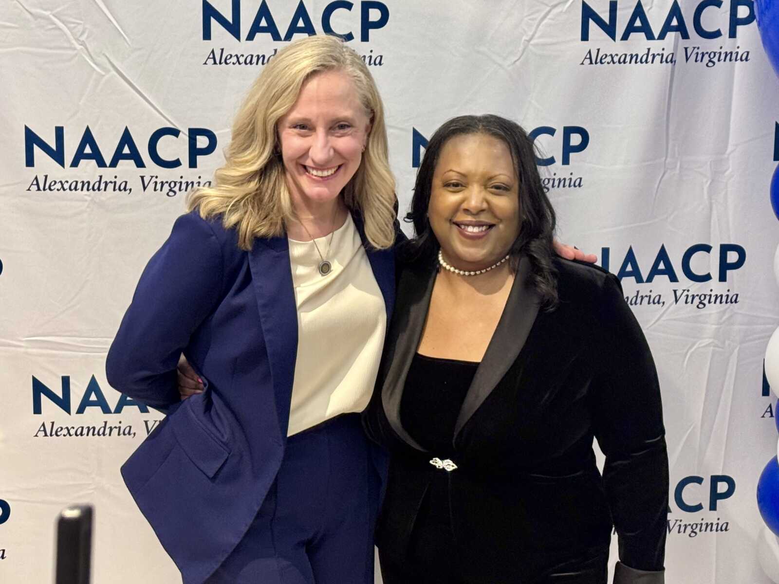 Two women smiling for a photo at a NAACP Alexandria, Virginia event, standing in front of a branded backdrop.