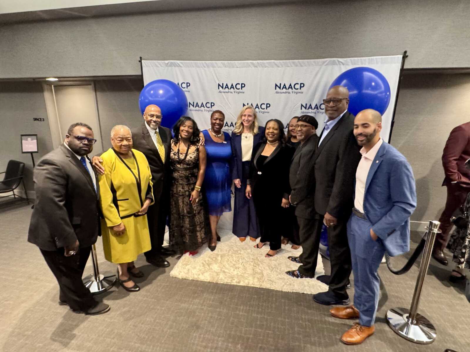 Eleven people in formal attire posing together at a NAACP event, with blue balloons and a NAACP backdrop in the background.
