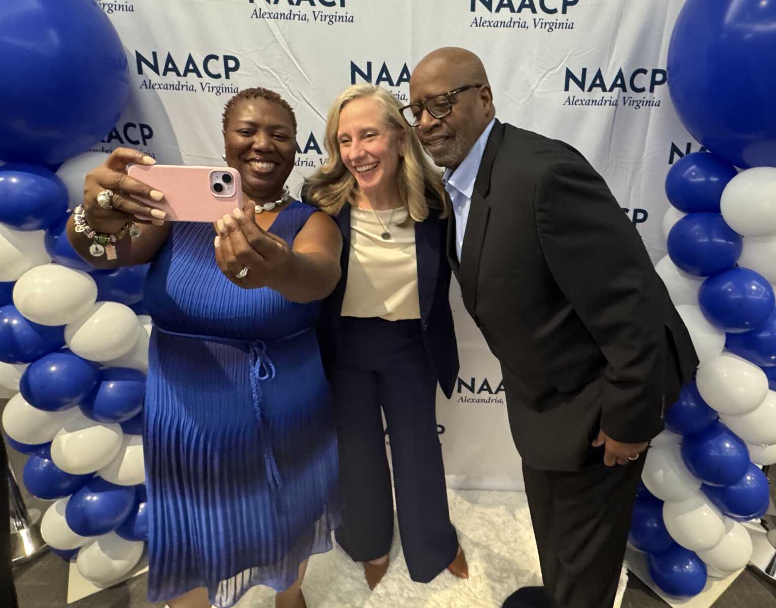 Three smiling adults pose for a group selfie at a NAACP Alexandria, Virginia event, with blue and white balloons in the background.