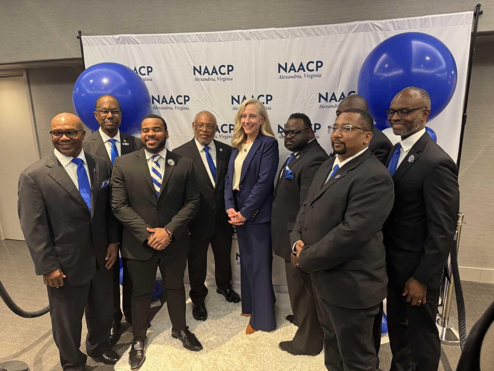 Group of professionally dressed individuals pose for a photo in front of a NAACP backdrop with blue balloons behind them, central woman in a blue suit.