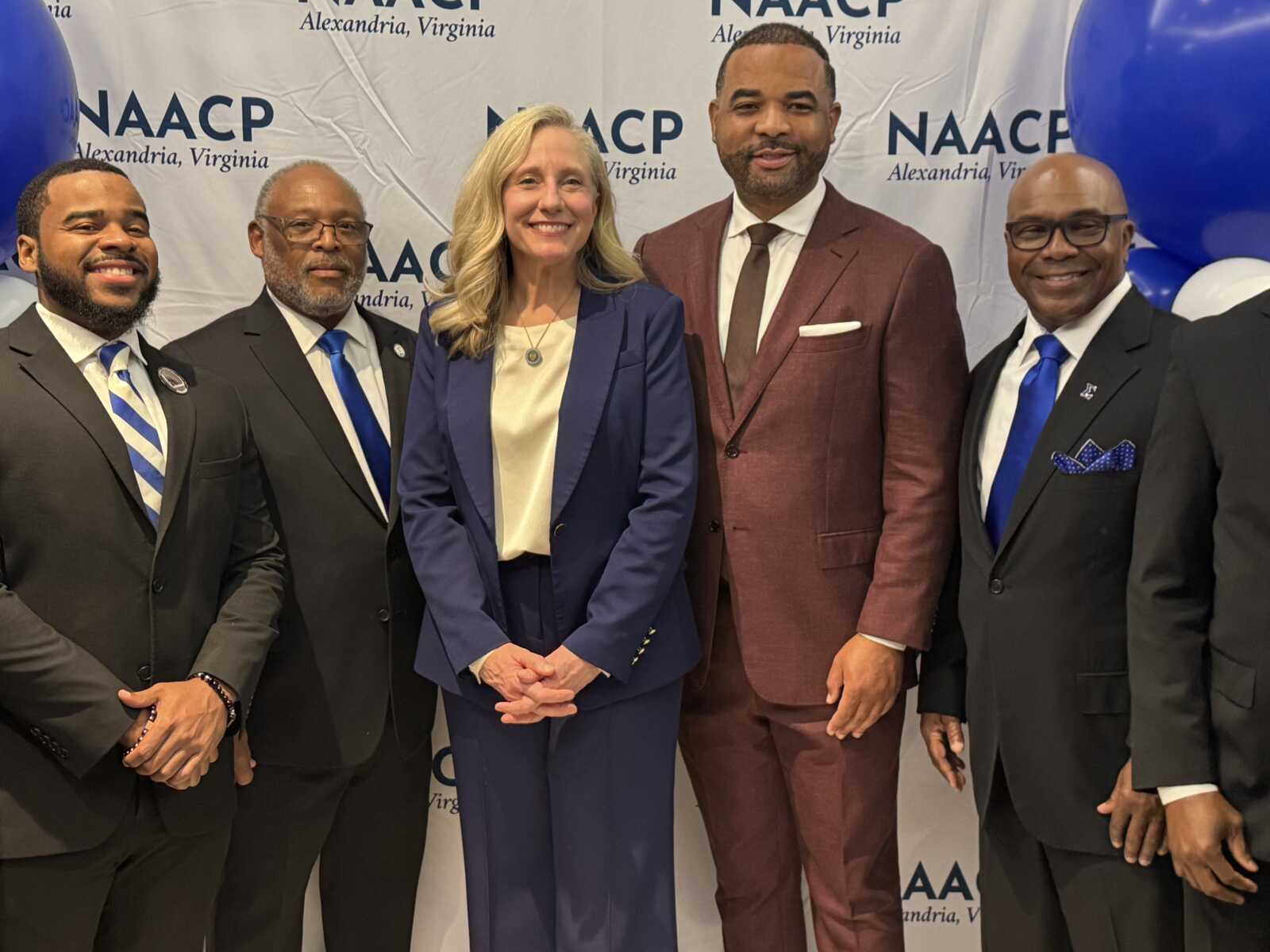 Five professionally dressed adults posing for a photo at a NAACP Alexandria event in front of a banner.0It features blue accents and balloons behind them.