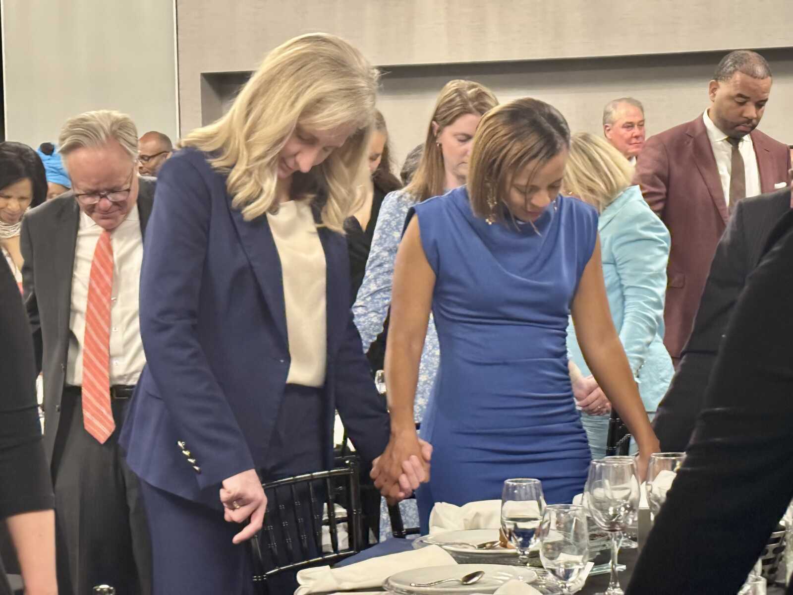 Group of formally dressed people standing and holding hands over a table set for dinner, heads bowed in a moment of prayer or reflection.