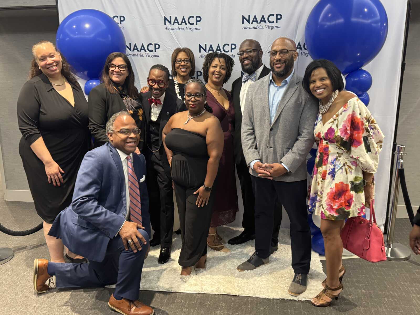 Group of ten professionally dressed adults posing together at a NAACP event, with blue balloons and a NAACP backdrop in the background.