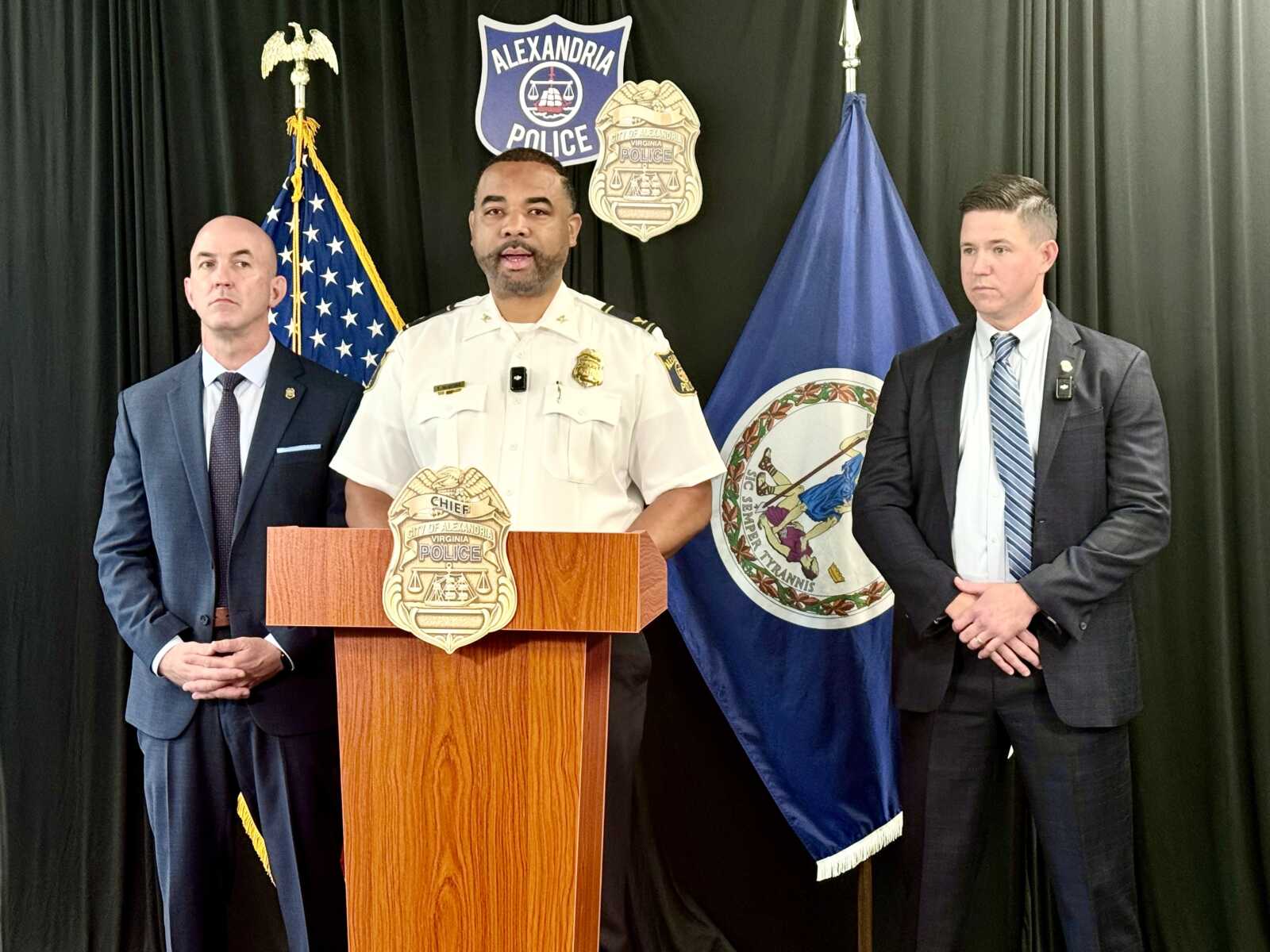 Three men stand at a wooden podium during a police press briefing: a uniformed chief speaking, flanked by two suited officers, with US and Virginia flags and police insignia in the background.
