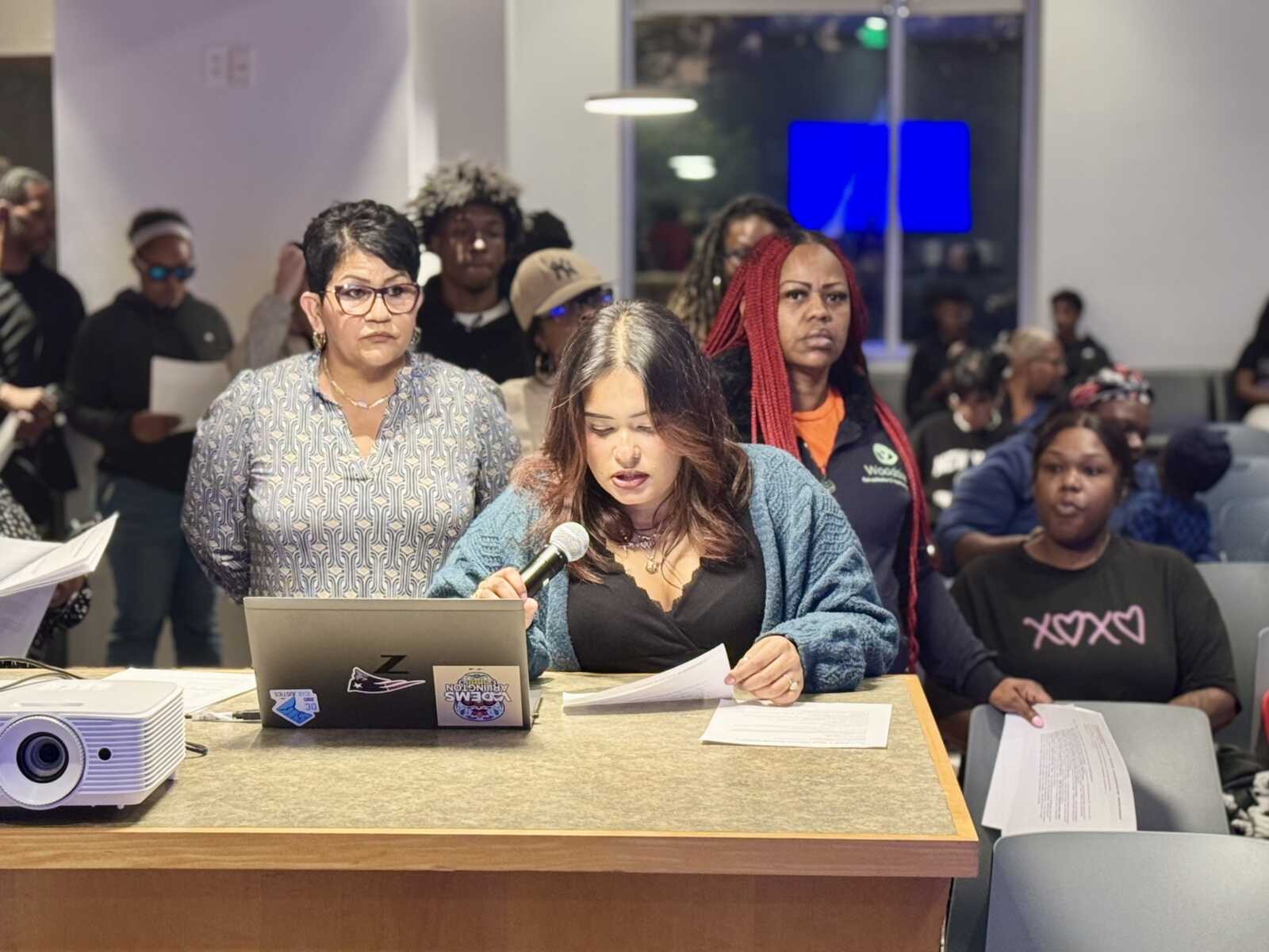 Woman in a blue cardigan speaks into a microphone at a desk with papers and laptop, while colleagues listen in a conference room.
