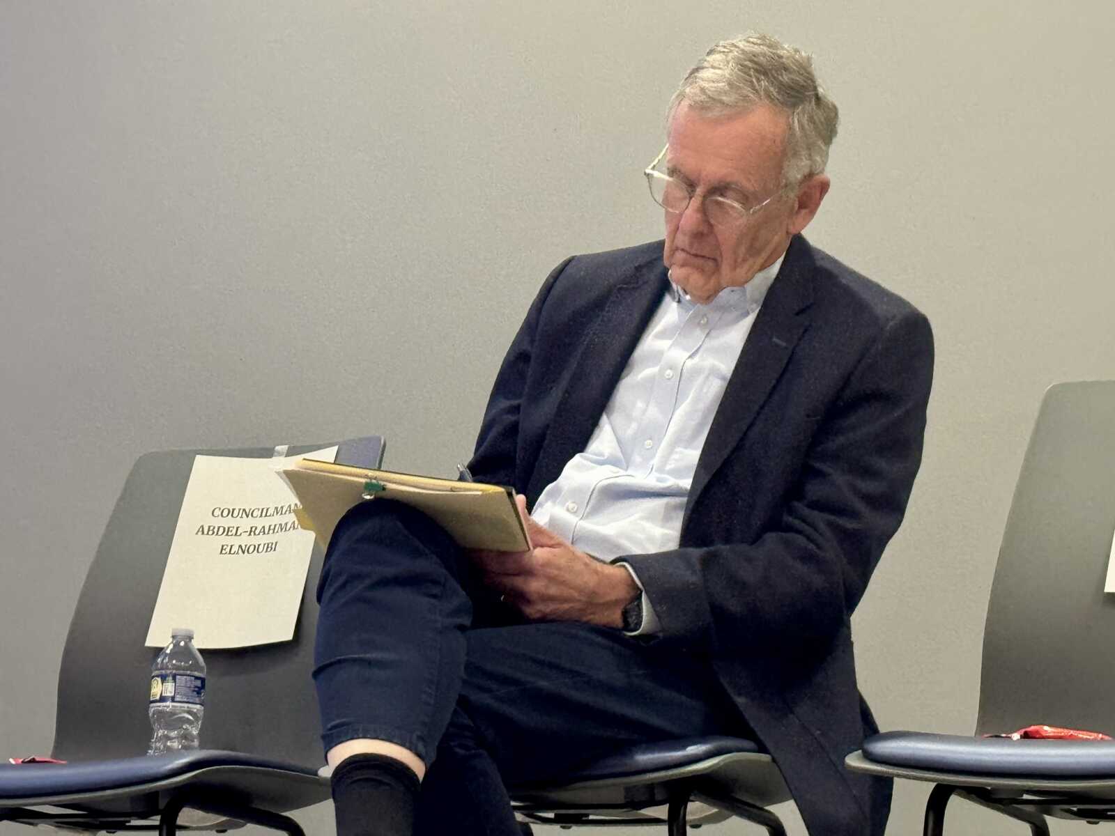 Older man with gray hair and glasses, wearing a dark blazer, reading a notebook while seated in a panel discussion room. A water bottle and documents are on the table nearby.